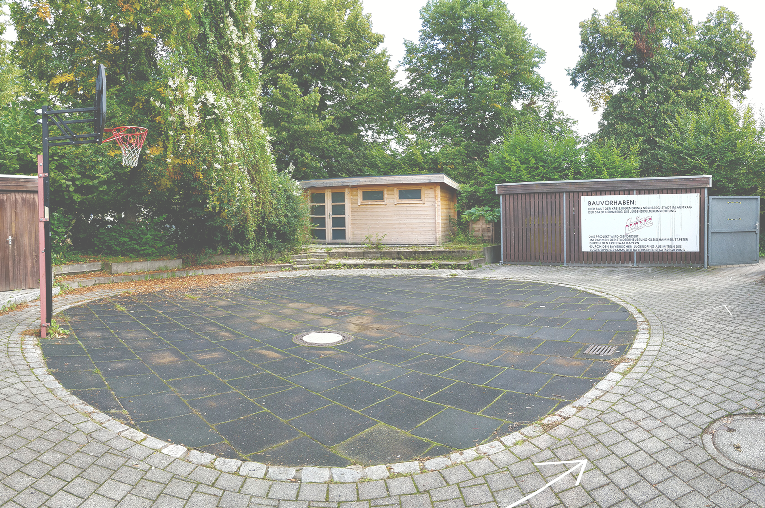 Foto des Außenbereiches mit rundem Basketballplatz und einem Baskettballkorb. um die runde Fläche herum drei Holzschuppen.